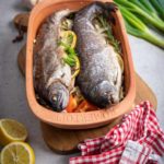 Zwei Forellen im Tontopf auf Gemüsebett mit Zitrone, Knoblauch und Frühlingszwiebeln, umgeben von Küchenutensilien.