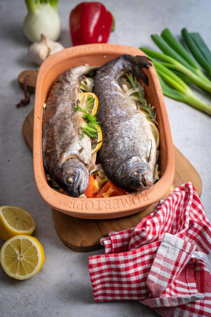 Zwei Forellen im Tontopf auf Gemüsebett mit Zitrone, Knoblauch und Frühlingszwiebeln, umgeben von Küchenutensilien.