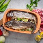 Zwei gebackene Forellen mit Zitrone und Kräutern in einer Römertopf-Form, umgeben von Gemüse auf einem Tisch.