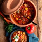 Ton-Topf mit herzhaftem Eintopf, serviert in Schüssel mit Sahne. Daneben rote Paprika, Karotten und Kräuter.