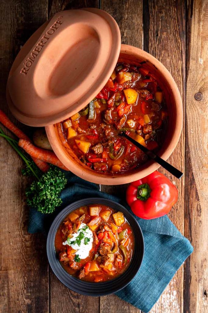 Ton-Topf mit herzhaftem Eintopf, serviert in Schüssel mit Sahne. Daneben rote Paprika, Karotten und Kräuter.