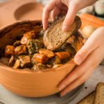 Hand tunkt Brot in einen Eintopf mit Gemüse und Fleisch in einem braunen Topf.