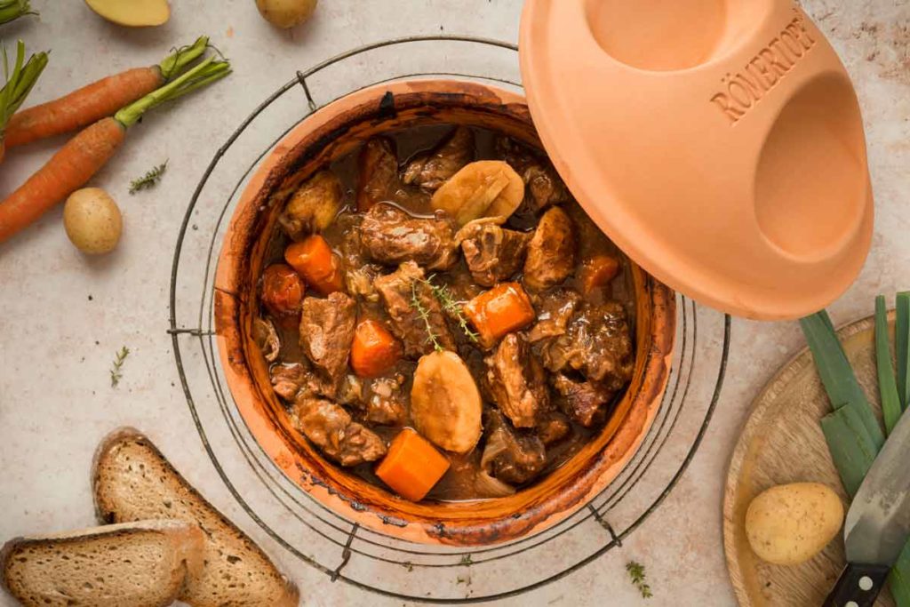 Eintopf im Römertopf mit Fleisch, Karotten und Kartoffeln, umgeben von frischem Gemüse und Brotscheiben.