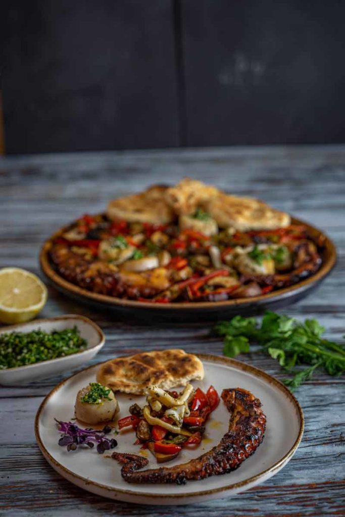 Teller mit gegrilltem Oktopus, Gemüse, Fladenbrot und Kräutern auf Holztisch, Zitronenscheiben und Koriander im Hintergrund.