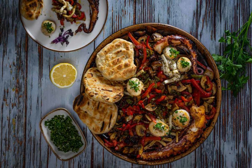 Gegrillter Pulpo mit Brot und Gemüse auf einem Teller, Zitrone und Kräuter auf Holztisch.