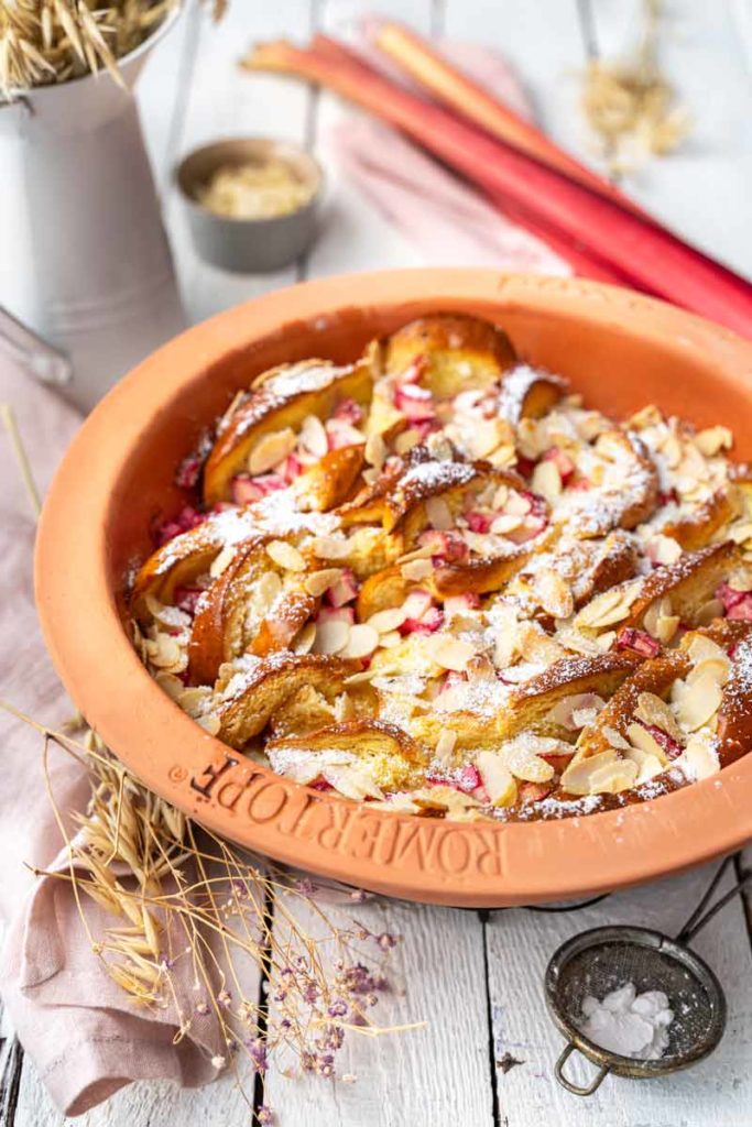Erdbeerauflauf mit gehobelten Mandeln in einer Tonform, dekoriert mit Puderzucker, auf hellen Holztisch fotografiert.