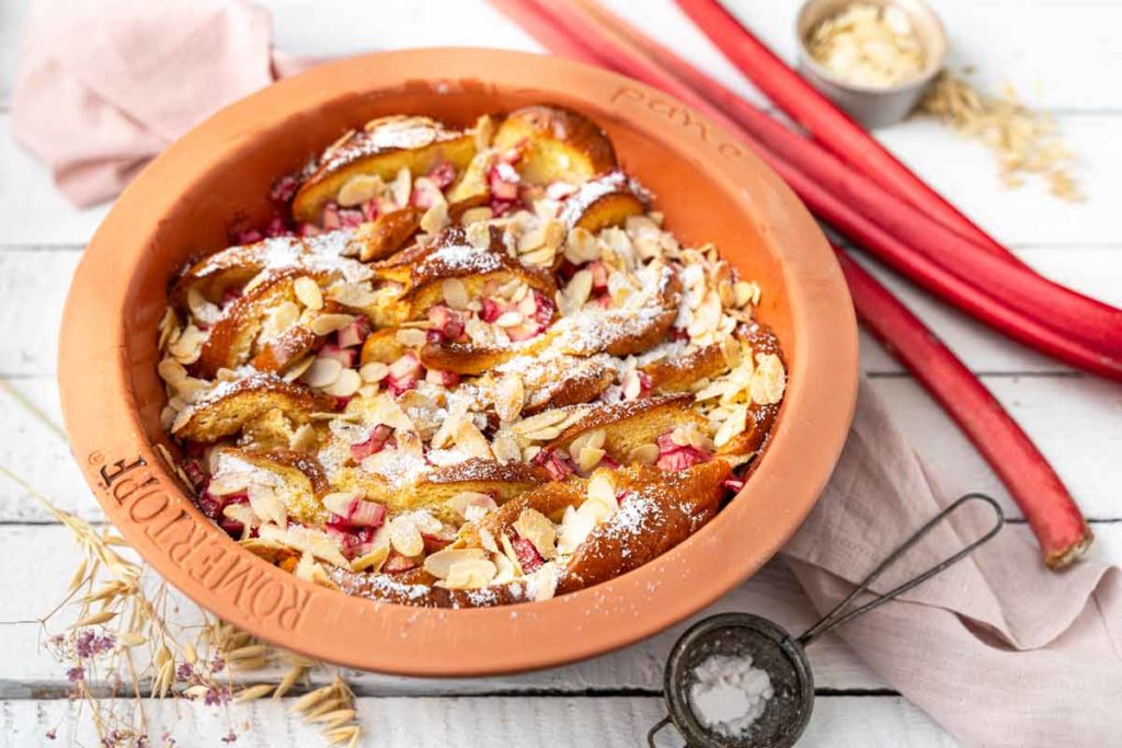 Rhabarber-Auflauf mit Mandeln in einer Tonform auf weißem Holztisch, dekoriert mit Hafer und Rhabarberstangen.
