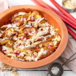 Rhabarber-Auflauf mit Mandeln in einer Tonform auf weißem Holztisch, dekoriert mit Hafer und Rhabarberstangen.