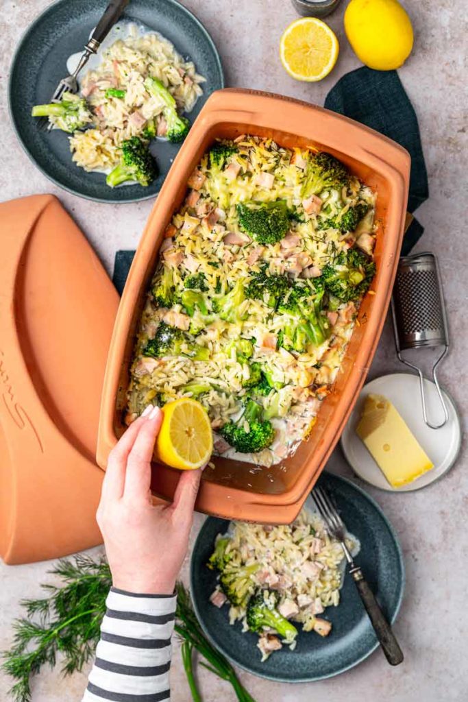 Hand presst Zitrone über Auflauf mit Brokkoli, Nudeln und Käse in einer Terrakottaform, daneben zwei Teller der Mahlzeit.