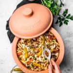 Römertopf mit Nudeln und Gemüse, daneben Petersilie, eingelegte Peperoni und eine Hand mit Löffel voller Gewürze.