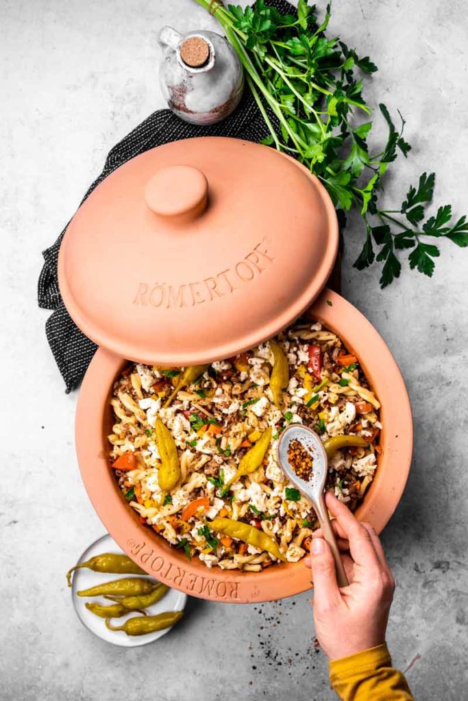 Römertopf mit Nudeln und Gemüse, daneben Petersilie, eingelegte Peperoni und eine Hand mit Löffel voller Gewürze.