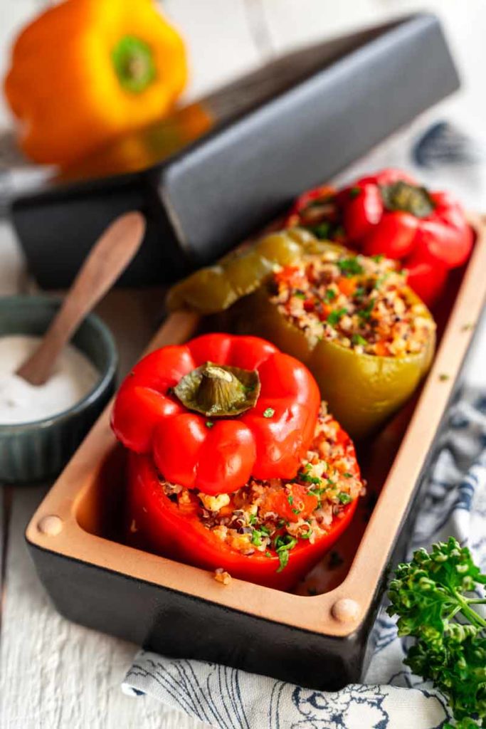Gefüllte Paprikaschoten mit Quinoa in einer Auflaufform, daneben eine Schüssel mit heller Sauce und ein Holzlöffel.