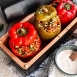 Drei gefüllte Paprikaschoten in einer Backform, mit einer Quinoa-Mischung, daneben eine Schale mit weißer Sauce.