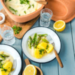 Teller mit Kartoffeln, Spargel und Sauce, serviert neben einem Römer-Topf, Zitronenhälften und Gläsern mit Wasser.