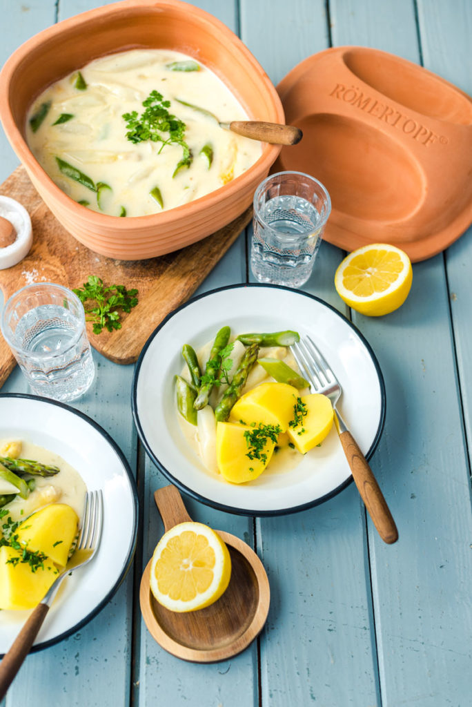 Teller mit Kartoffeln, Spargel und Sauce, serviert neben einem Römer-Topf, Zitronenhälften und Gläsern mit Wasser.