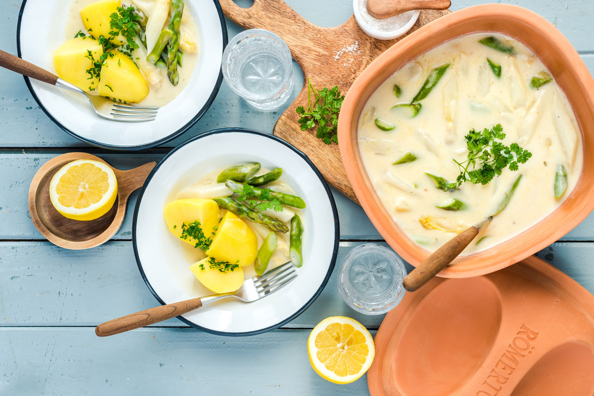 Zwei Teller mit Spargel, Kartoffeln und Soße. Im Hintergrund ein Tontopf mit Spargelragout, Zitrone und Petersilie.