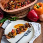 Teller mit gegrilltem Gemüsespieß und einer Gabel, im Hintergrund ein gebackenes Gericht und Paprika.