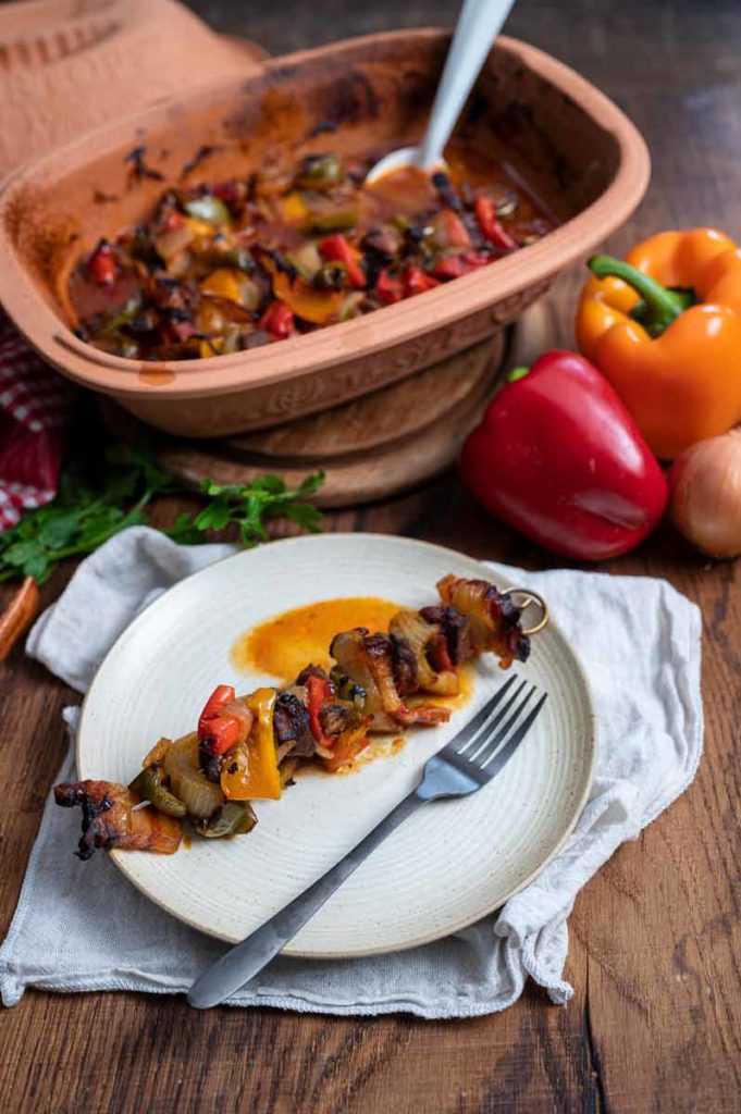 Teller mit gegrilltem Gemüsespieß und einer Gabel, im Hintergrund ein gebackenes Gericht und Paprika.