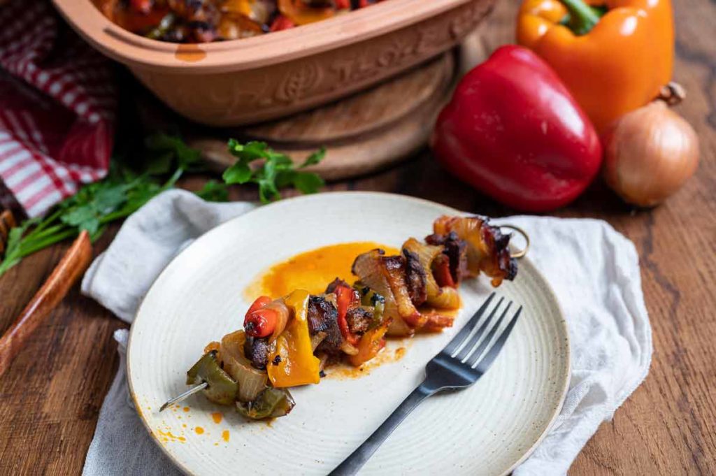 Gegrillter Gemüsespieß auf einem Teller, umgeben von Paprika und Zwiebeln; eine Gabel liegt daneben.