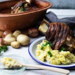 Gebackene Schweinshaxe mit Kartoffelpüree auf Teller, umgeben von rohen Kartoffeln und Tontopf im Hintergrund.