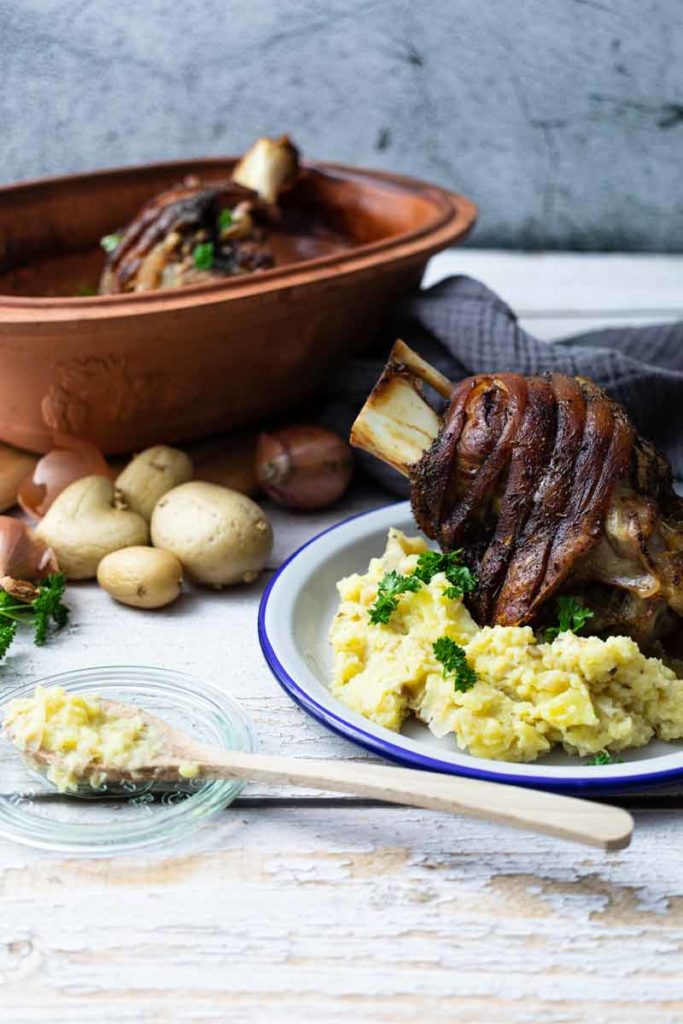 Gebackene Schweinshaxe mit Kartoffelpüree auf Teller, umgeben von rohen Kartoffeln und Tontopf im Hintergrund.