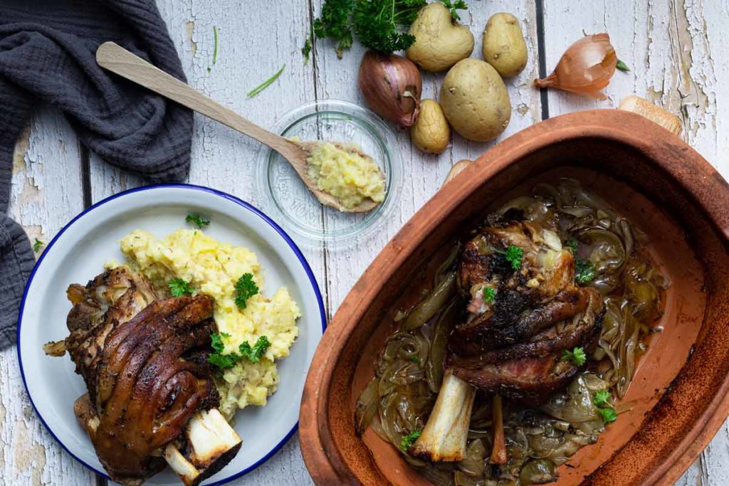 Gebackene Schweinshaxe mit Kartoffelpüree auf einem Teller; daneben Zutaten und eine Auflaufform mit Zwiebeln.