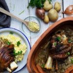 Gebackene Schweinshaxe mit Kartoffelpüree auf einem Teller; daneben Zutaten und eine Auflaufform mit Zwiebeln.