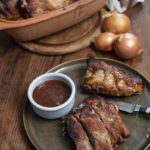 Gebratene Rippchen auf einem Teller mit BBQ-Sauce, Tonbräter im Hintergrund und Zwiebeln daneben.