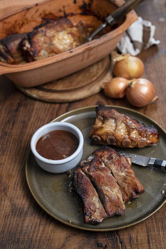 Gebratene Rippchen auf einem Teller mit BBQ-Sauce, Tonbräter im Hintergrund und Zwiebeln daneben.