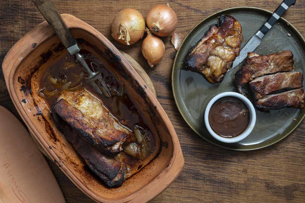 Geröstete Rippchen in einer Tonform neben einem Teller mit Rippchenteilen, Zwiebeln und Barbecuesoße.