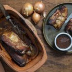 Geröstete Rippchen in einer Tonform neben einem Teller mit Rippchenteilen, Zwiebeln und Barbecuesoße.