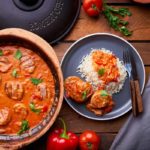 Römertopf mit Gulasch, serviert mit Reis, Paprika und Tomaten auf einem Teller, daneben Besteck.