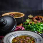 Teller mit Erbsensuppe, Wurst und Speck, im Hintergrund frische Kräuter und ein Kochtopf mit geschlossener Kasserollenabdeckung.