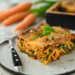 Stück Gemüse-Lasagne mit Karotten und Spinat auf einem Teller, garniert mit Petersilie, daneben eine Gabel.