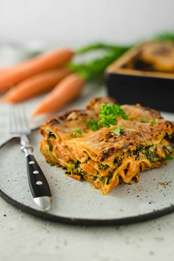 Stück Gemüse-Lasagne mit Karotten und Spinat auf einem Teller, garniert mit Petersilie, daneben eine Gabel.