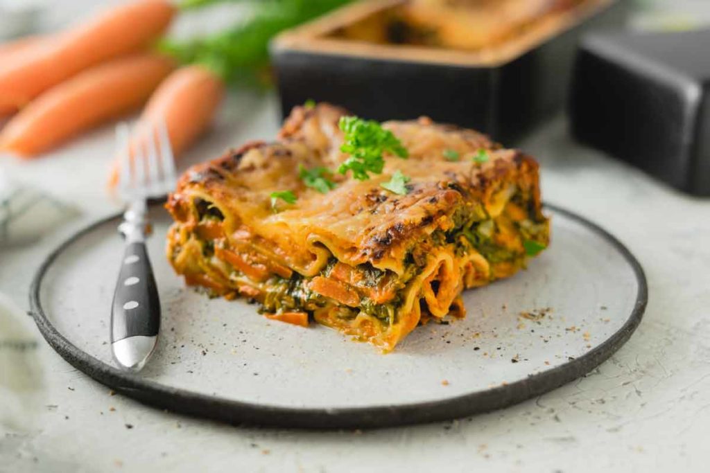 Eine Scheibe Gemüse-Lasagne auf einem Teller, garniert mit Kräutern, daneben eine Gabel. Im Hintergrund liegen Karotten.