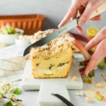 Person schneidet Rhabarberkuchen mit Streuseln auf einem weißen Brett. Zutaten und Blumen liegen im Hintergrund.