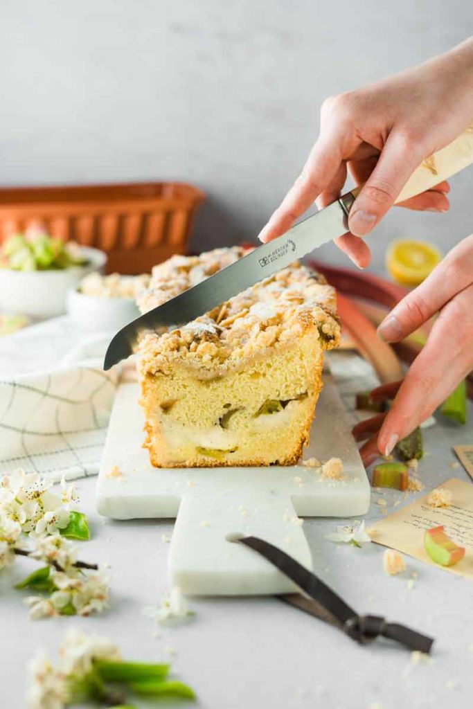 Person schneidet Rhabarberkuchen mit Streuseln auf einem weißen Brett. Zutaten und Blumen liegen im Hintergrund.
