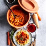 Römertopf mit geschmortem Fleisch und Gemüse, Nudeln, Wein und Gewürze auf einem Tisch arrangiert.