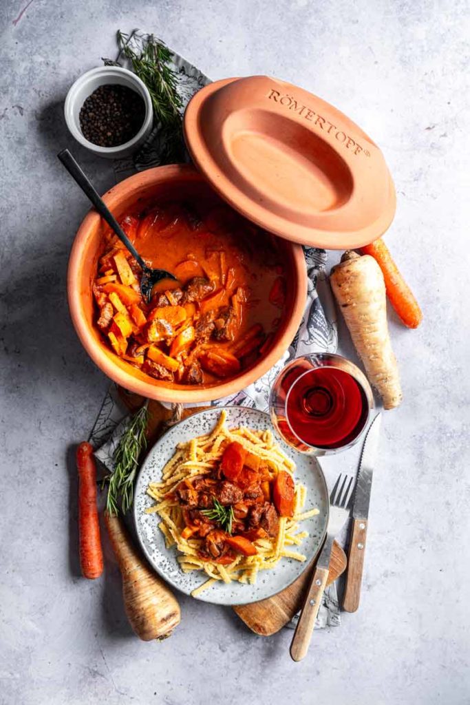 Römertopf mit geschmortem Fleisch und Gemüse, Nudeln, Wein und Gewürze auf einem Tisch arrangiert.