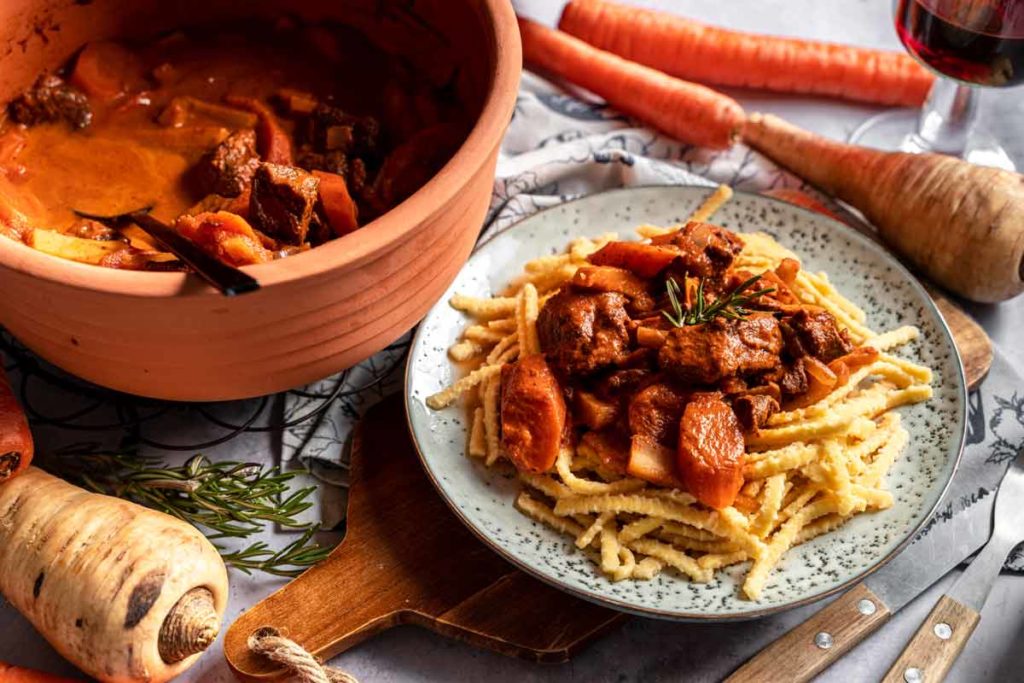 Teller mit Spätzle und Rindergulasch, garniert mit Rosmarin, daneben ein Topf mit Gulasch, Karotten und Pastinaken.