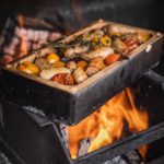 Bratwürste und Tomaten in einem Ofen auf offenem Feuer im Backblech garen, mit Kräuterzweig garniert.