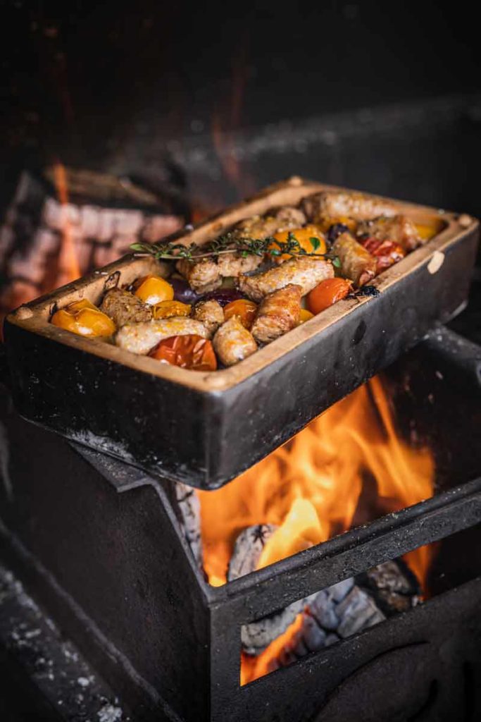 Bratwürste und Tomaten in einem Ofen auf offenem Feuer im Backblech garen, mit Kräuterzweig garniert.