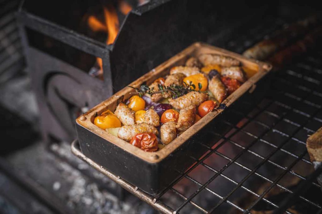 Bratwürste und Kirschtomaten in einer rechteckigen Form auf einem Grill über offenem Feuer gekocht.