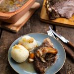 Teller mit Braten und zwei Knödeln, garniert mit Zwiebeln, auf rustikalem Holztisch, Bräter und Besteck daneben.