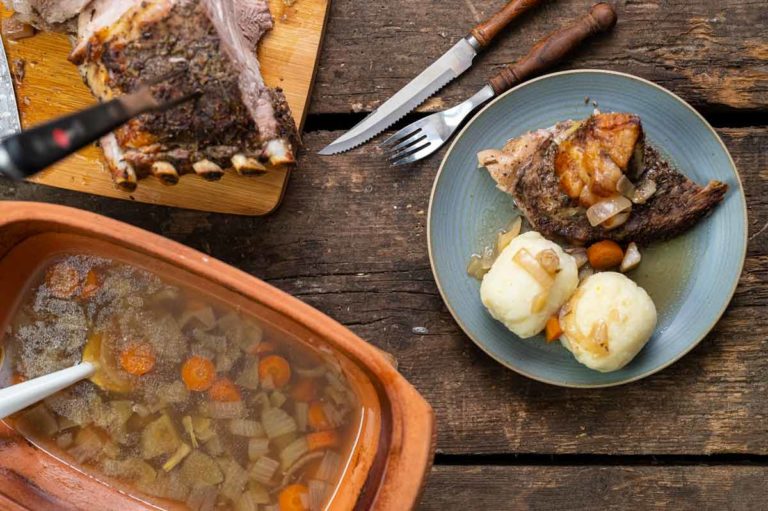 Teller mit Braten und Knödeln, daneben Brühe in einem Topf und geschnittenes Fleisch auf Holzbrett auf rustikalem Tisch.