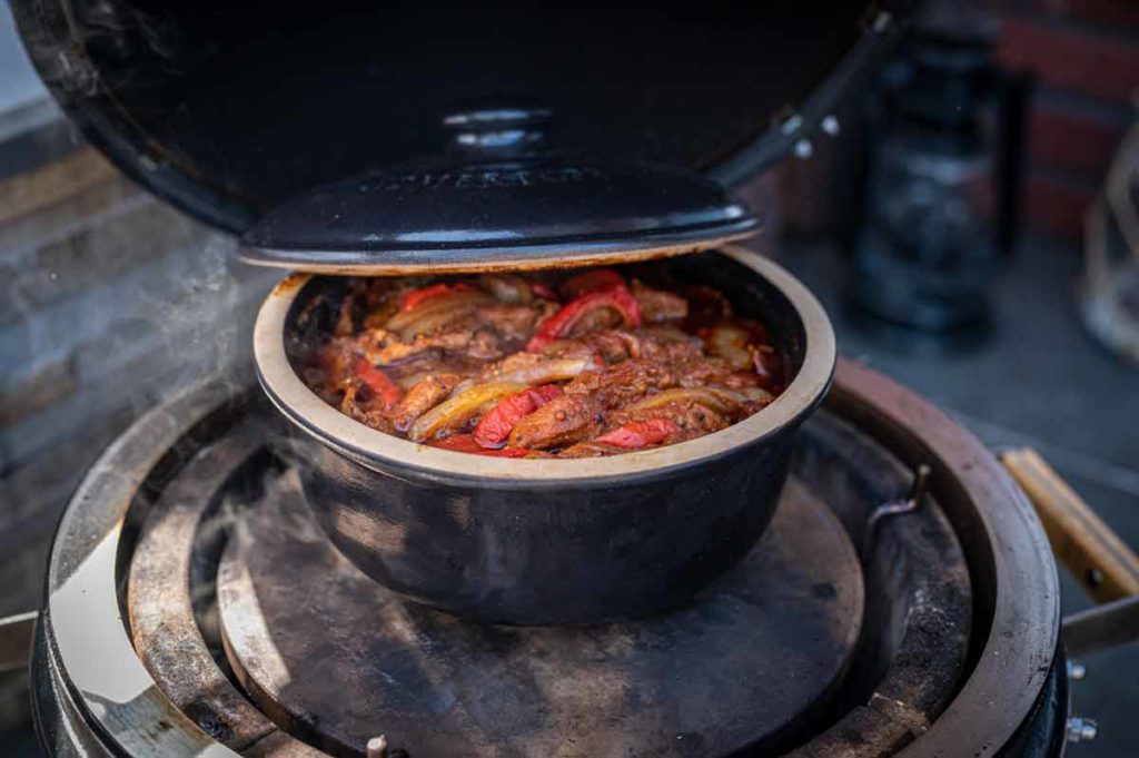 Schmortopf mit Gemüse und Fleisch auf einem Grill, mit teilweise geöffnetem Deckel.
