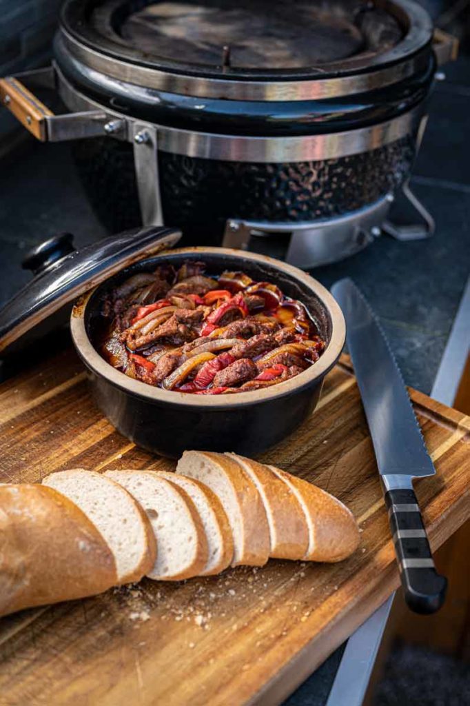 Eintopf mit Paprika und Fleisch in einem Topf auf Holzbrett, daneben geschnittenes Brot und ein Messer.