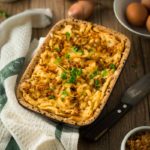 Auflaufform mit Kässpätzle, garniert mit Petersilie, auf Holztisch. Daneben Küchentuch, Messer und Zutaten wie Eier.