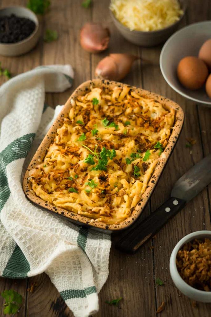 Auflaufform mit Kässpätzle, garniert mit Petersilie, auf Holztisch. Daneben Küchentuch, Messer und Zutaten wie Eier.